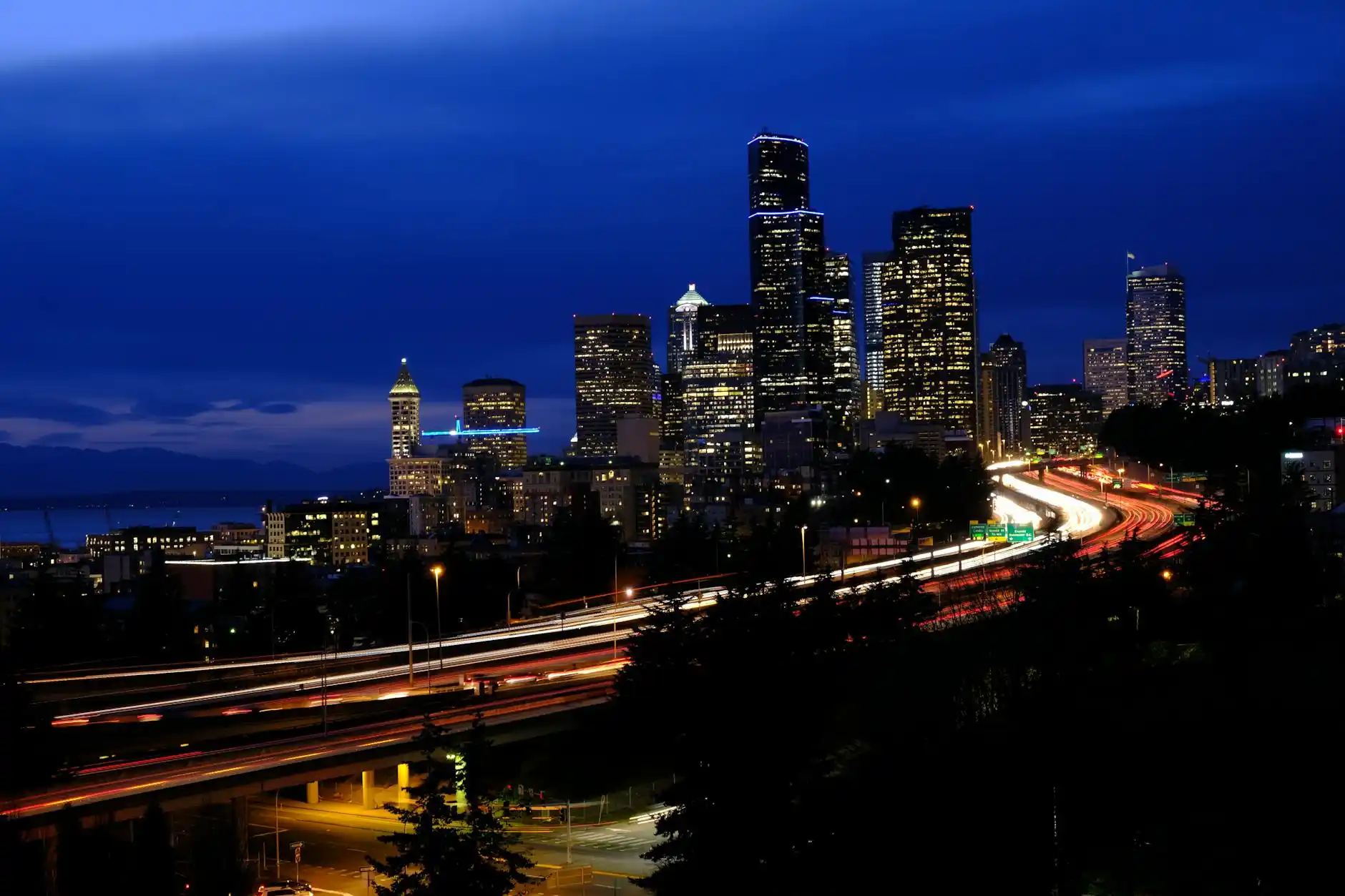 sleek black luxury limousine parked curbside in downtown Seattle at dusk, glowing city lights, Space Needle softly visible in background, chauffeur opening door for elegantly dr...