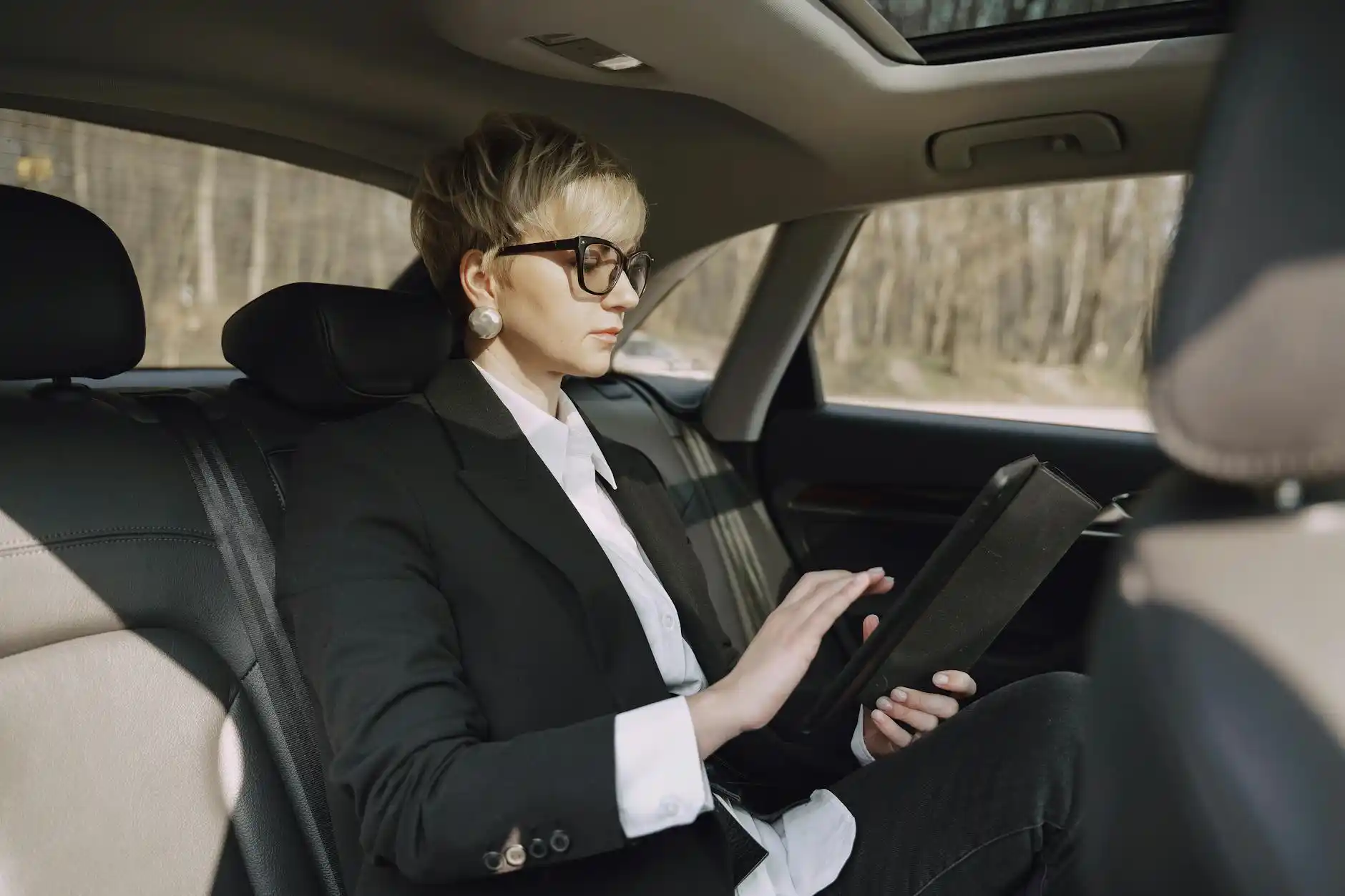elegantly dressed business traveler seated in rear of luxury sedan using tablet, rain-slick Seattle streets outside, modern office towers and traffic blurred in background